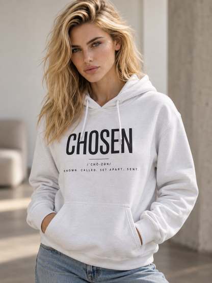 A blonde woman wearing a white pullover hoodie with 'CHOSEN' printed in bold black letters on the front, paired with light blue jeans, in a brightly lit indoor setting.