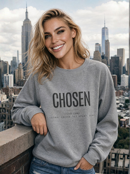 A smiling blonde woman wearing a heather gray crewneck sweatshirt with 'CHOSEN' printed in black across the chest, posed outdoors against a city skyline background.