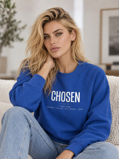 A blonde woman sitting on a couch wearing a royal blue crewneck sweatshirt with 'CHOSEN' printed in white across the chest, paired with light blue jeans.