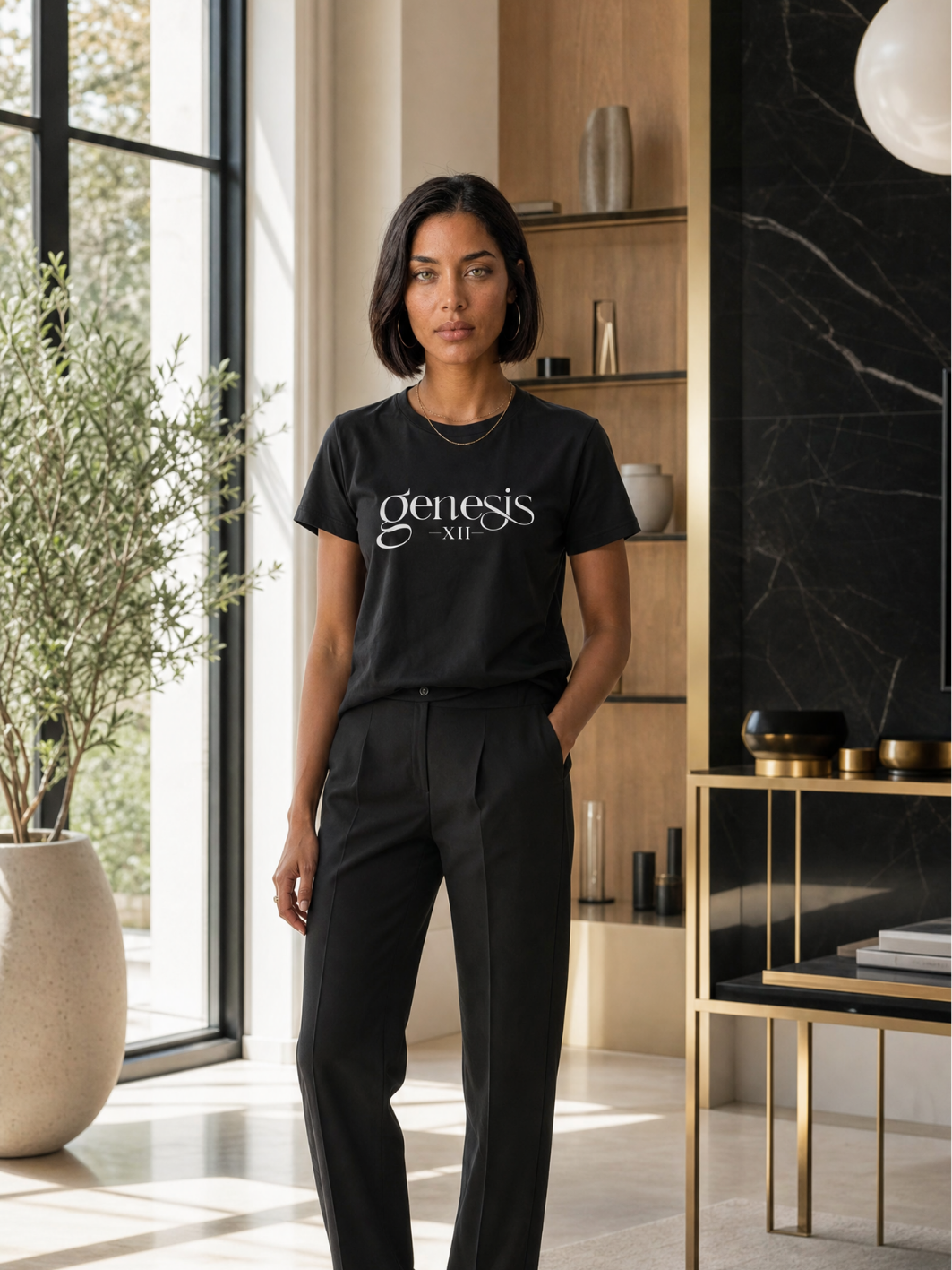 A woman stands in a modern interior wearing a black short-sleeve T-shirt with the white script text 'genesis XII' across the chest, paired with black trousers.
