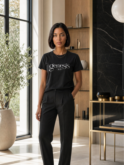 A woman stands in a modern interior wearing a black short-sleeve T-shirt with the white script text 'genesis XII' across the chest, paired with black trousers.