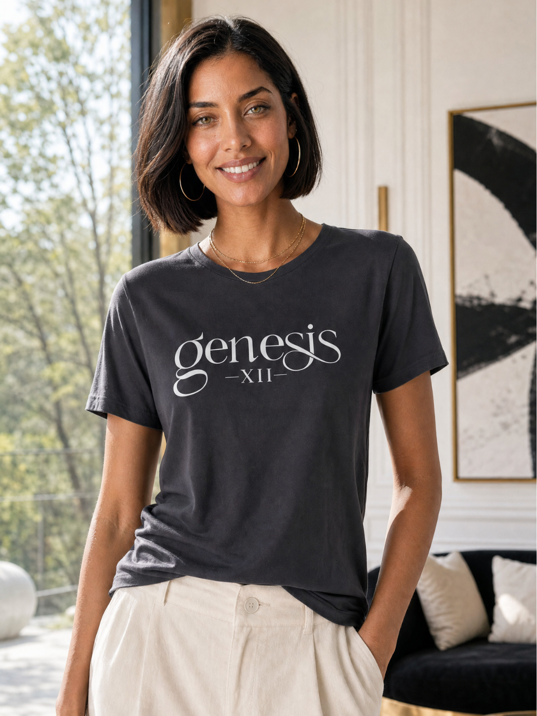A woman wearing a dark charcoal gray t-shirt with 'genesis XII' printed in white script, paired with cream-colored pants, standing in a bright, modern room.