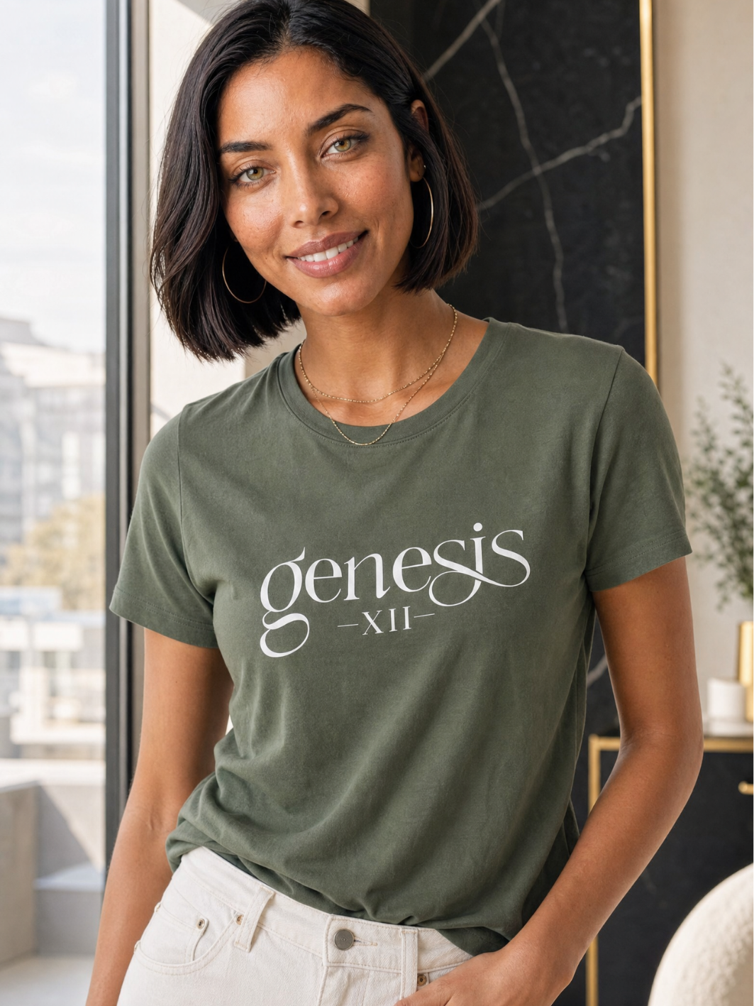 A woman wearing an olive green / military green t-shirt with 'genesis XII' printed in white script on the front, paired with white pants, standing in a modern indoor setting.