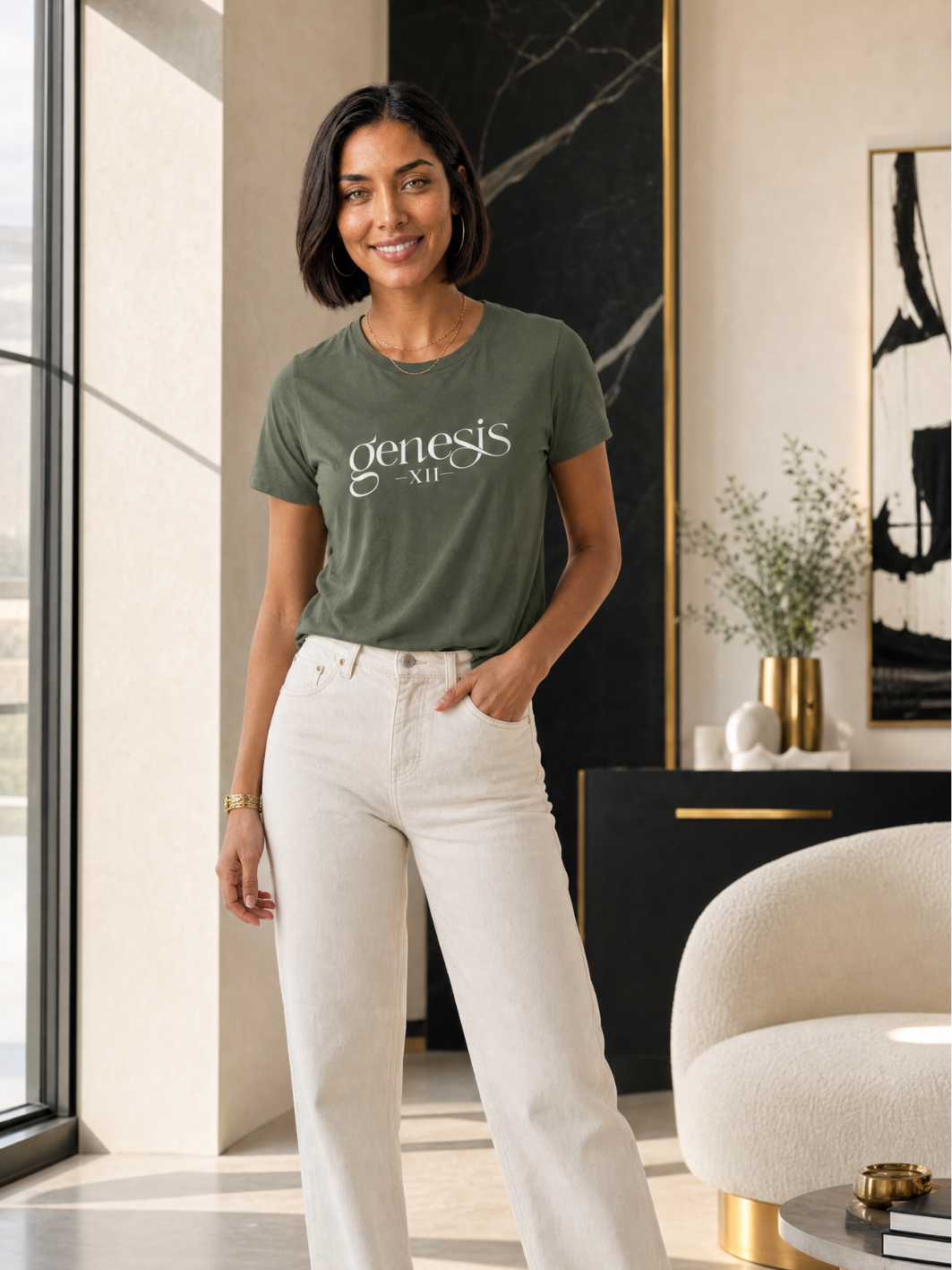 A woman stands in a modern interior wearing an olive / militar green graphic T-shirt with 'genesis AIR' printed in white across the chest, paired with off-white high-waisted jeans.