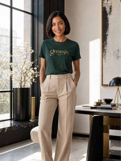 A woman stands in a modern room wearing a Forest Green green t-shirt with the word 'genesis' printed in gold on the chest, paired with beige high-waisted trousers.