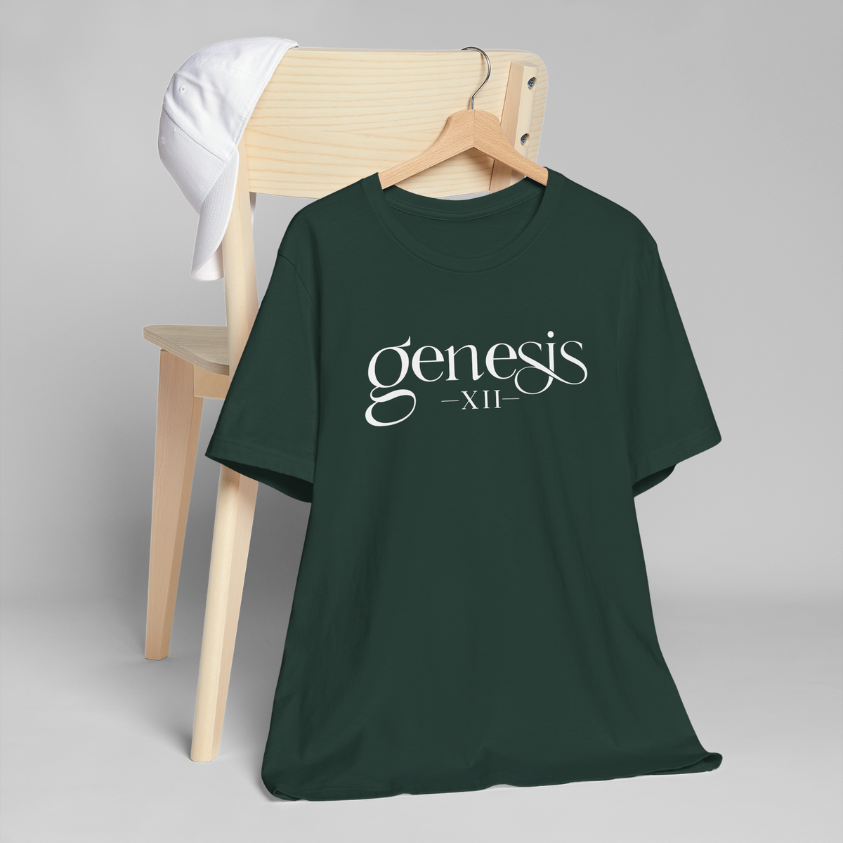 A dark forest green t-shirt with 'genesis XII' printed in white script across the chest, hanging on a wooden hanger from a light wood chair with a white shirt draped over it, set against a neutral gray background.